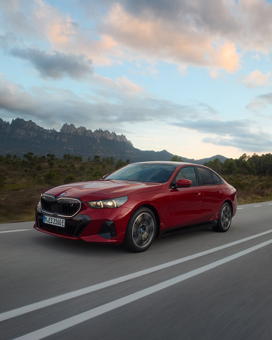 Ein roter BMW i5 Limousine fährt auf einer Landstraße bei Tageslicht, umgeben von hügeliger Landschaft und Nadelbäumen. Im Hintergrund sind markante Felsformationen und ein teilweise bewölkter Himmel zu sehen.