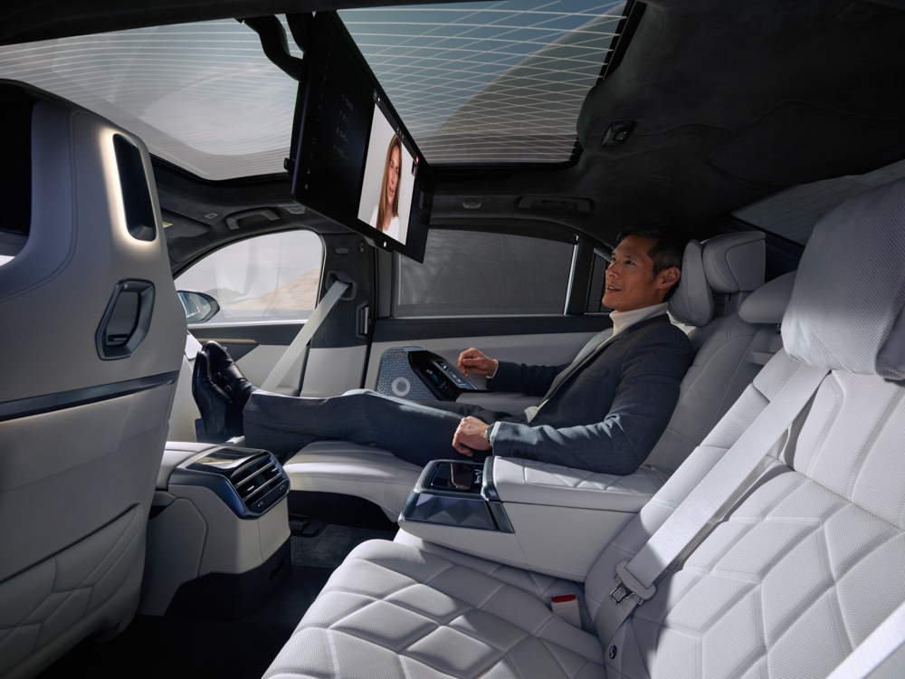 Ein Mann im Anzug lehnt sich auf dem Rücksitz eines modernen Autos mit weißer Lederausstattung zurück und schaut auf einen an der Decke montierten Bildschirm. Das Auto hat ein Panoramaglasdach und ein futuristisches Hightech-Design.