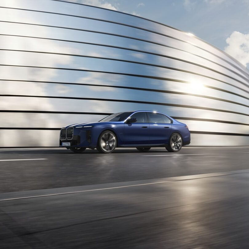 Eine blaue Limousine fährt auf einer gepflasterten Straße neben einem modernen, geschwungenen Metallgebäude mit horizontalen Linien, unter einem teilweise bewölkten Himmel.