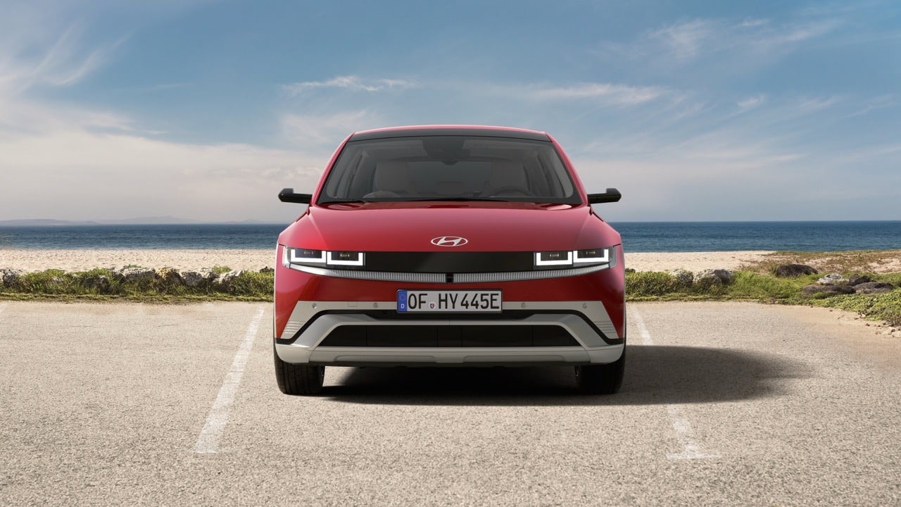 Ein rotes Hyundai Ioniq 5 Elektroauto steht mit dem Gesicht nach vorne auf einem Parkplatz in der Nähe eines Strandes, mit dem Meer und dem blauen Himmel im Hintergrund.
