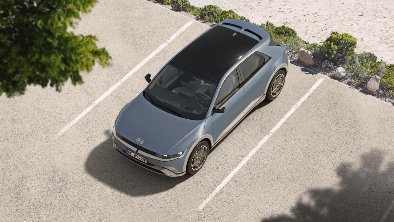 Ein silbernes Hyundai Ioniq 5-Elektroauto steht auf einem leeren Parkplatz in der Nähe einer sandigen Fläche mit Büschen und Steinen. Das Auto wird schräg von oben betrachtet.