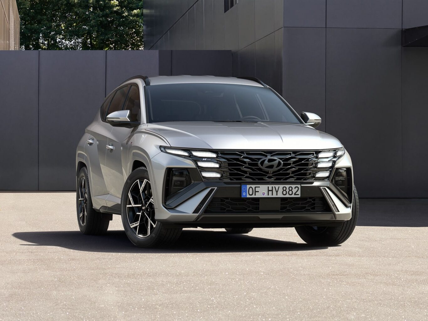 Ein silberner Hyundai-Geländewagen ist auf einer glatten, gepflasterten Fläche vor einem modernen Gebäude mit grauen Wänden und Glasscheiben geparkt. Die Frontpartie und das Nummernschild des Fahrzeugs sind sichtbar.