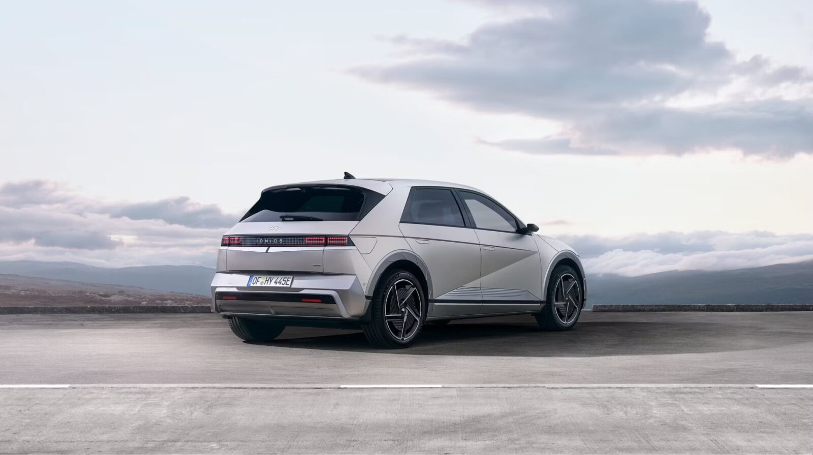 Ein silbernes Hyundai Ioniq 5 Elektroauto ist auf einer Betonfläche mit Bergen und bewölktem Himmel im Hintergrund geparkt.