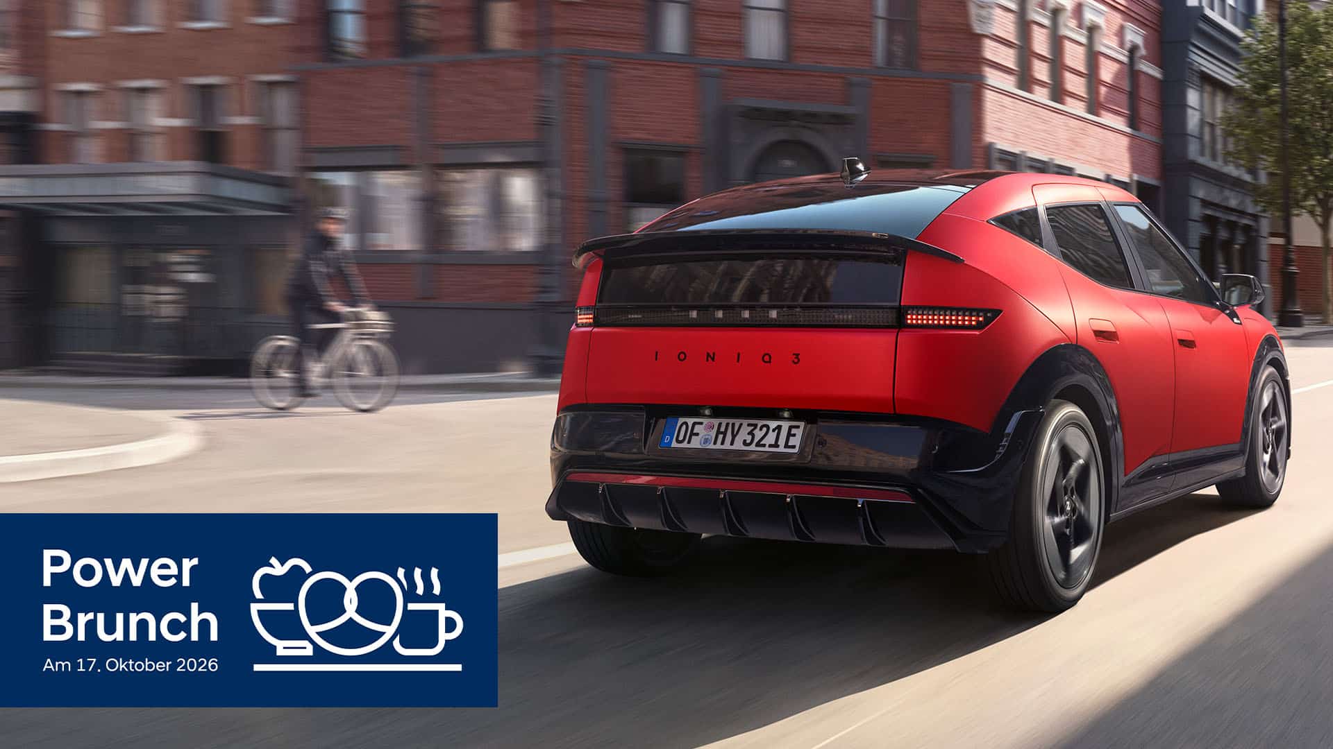 Ein roter Hyundai IONIQ 3 fährt auf einer Stadtstraße in der Nähe von Backsteinhäusern. Ein Radfahrer fährt auf dem Bürgersteig. Auf einem blauen Banner unten links steht Power Brunch am 17. Oktober 2026, begleitet von Symbolen für einen Stecker, eine Tasse und ein Croissant.