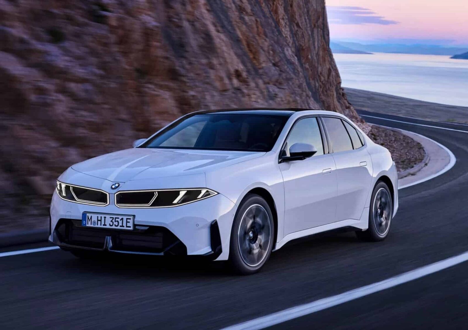 Eine weiße BMW i3 Limousine fährt bei Sonnenuntergang auf einer kurvenreichen Küstenstraße entlang einer felsigen Klippe, im Hintergrund sind das Meer und ein pastellfarbener Himmel zu sehen.