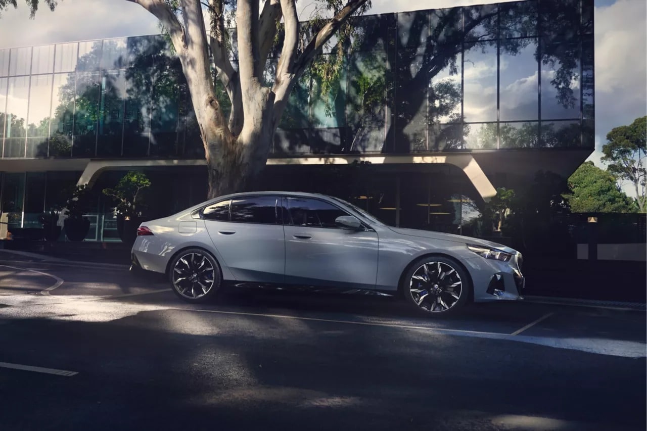 Eine silberne BMW i5 Limousine parkt auf einer Straße vor einem modernen Glasgebäude mit großen Bäumen, die Schatten auf die Szene werfen.
