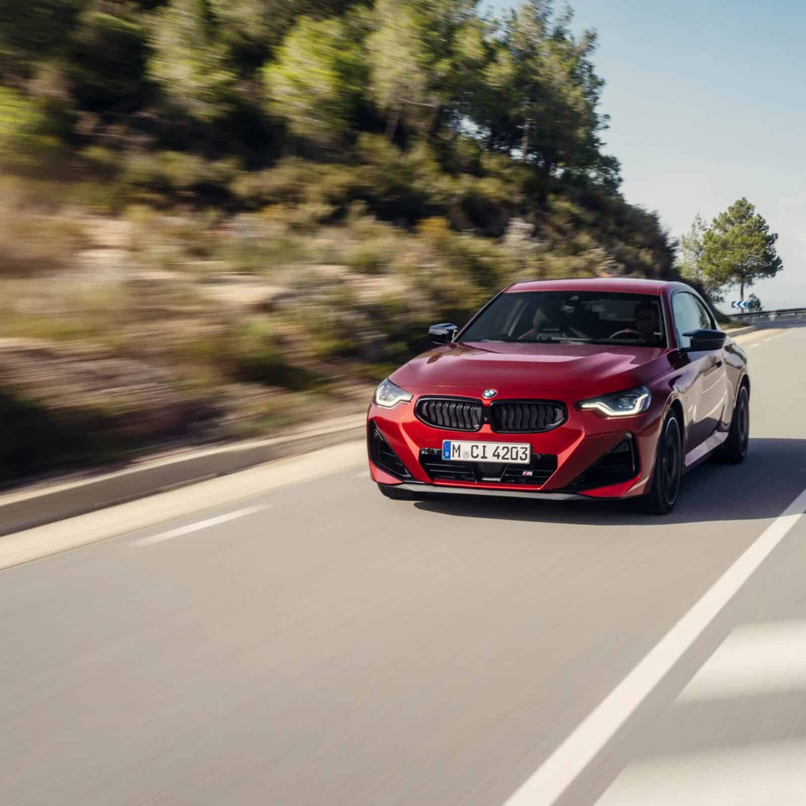 Ein rotes BMW 2er Coupé fährt auf einer zweispurigen Straße, die von Bäumen und Grünanlagen gesäumt ist, mit einem klaren Himmel im Hintergrund. Das Auto ist in Bewegung, und das Foto wurde von der vorderen linken Seite aufgenommen.
