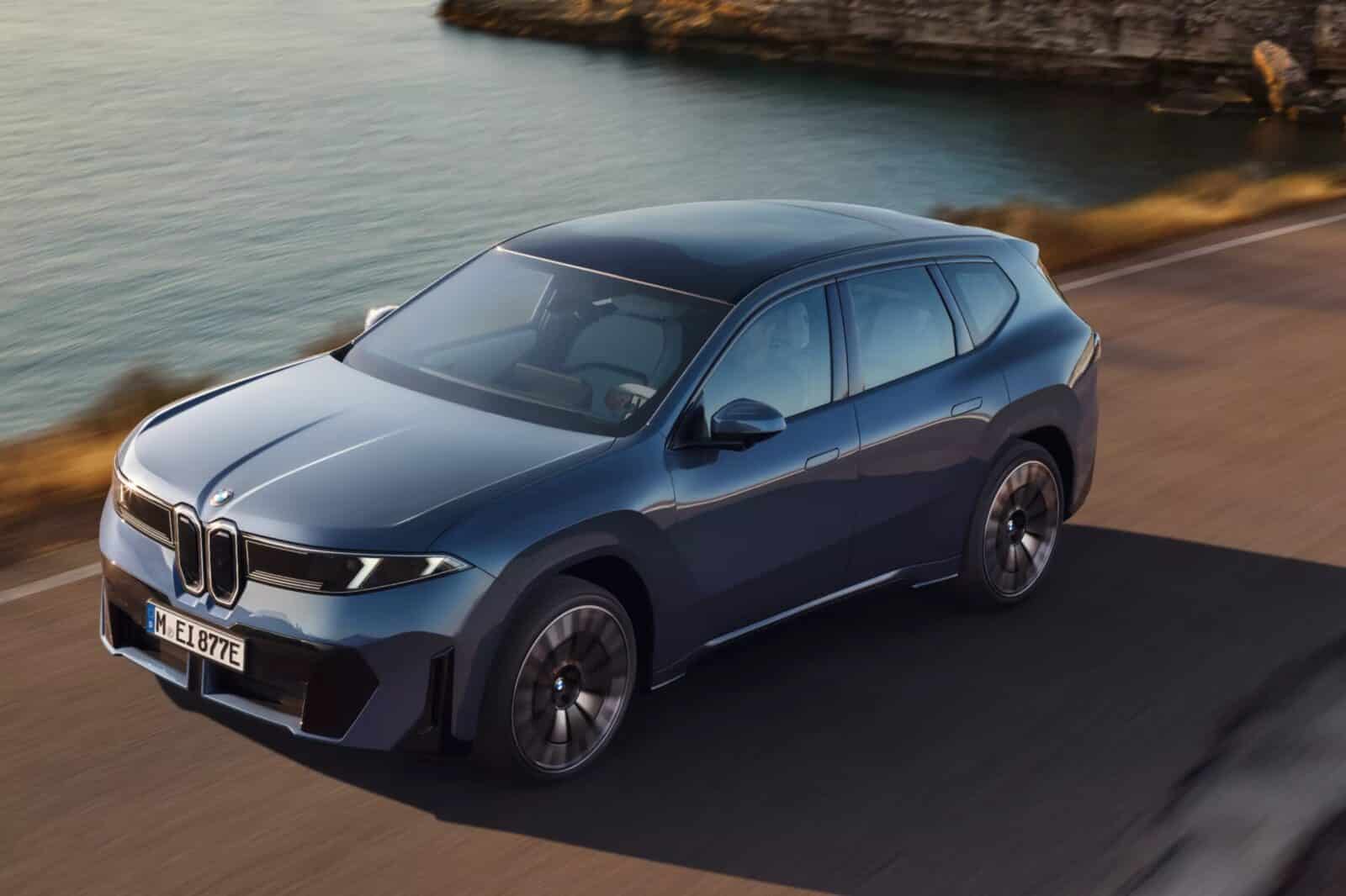 Ein blauer BMW iX3 fährt auf einer asphaltierten Küstenstraße neben einem Gewässer, wobei im Hintergrund felsige Klippen und trockenes Gras zu sehen sind. Das Foto ist aus einem erhöhten, frontalen Winkel aufgenommen.