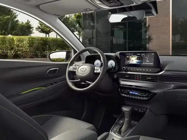 Das Bild zeigt den Innenraum eines modernen Autos mit schwarzem Armaturenbrett, Lenkrad, Touchscreen-Display, Schalthebel und Vordersitzen. An der Tür sind grüne Akzente zu sehen. Vor dem Fenster sind Bäume und ein Gebäude zu sehen.
