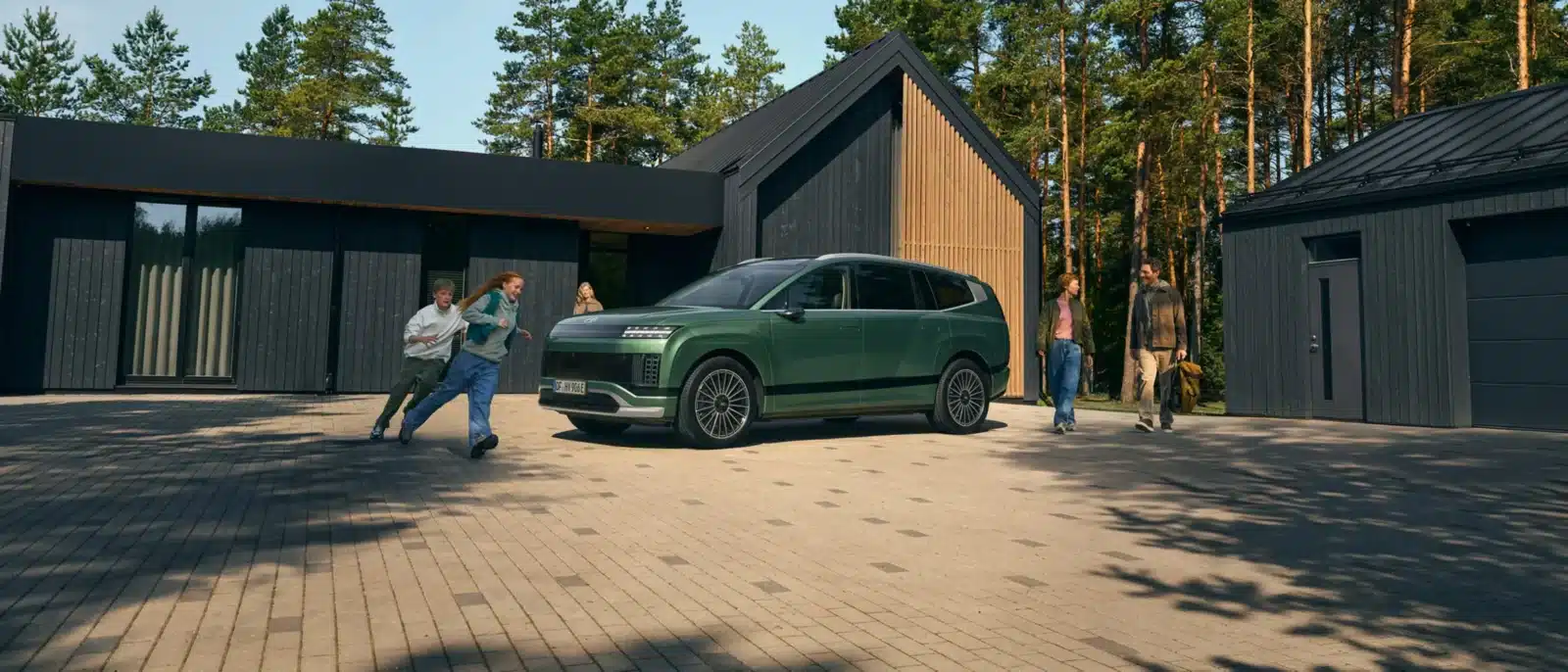 Drei Kinder spielen in der Nähe eines grünen Geländewagens, der vor einem modernen Haus mit Holz und dunkler Verkleidung geparkt ist. Zwei Erwachsene stehen in der Nähe, alle umgeben von Bäumen und einer gepflasterten Einfahrt.