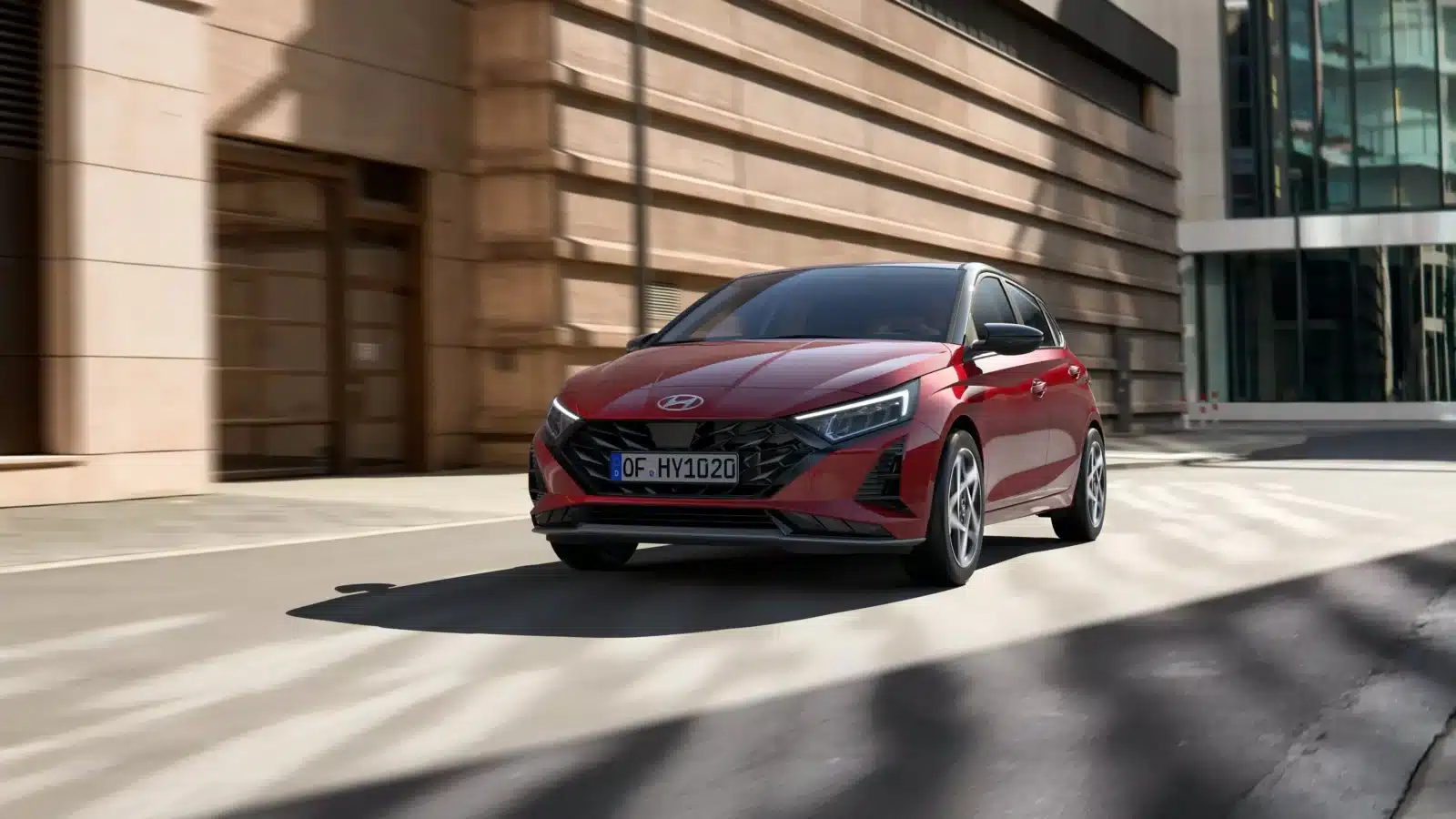 Ein rotes Hyundai-Auto fährt auf einer Stadtstraße mit modernen Gebäuden im Hintergrund. Das Auto ist schräg von vorne fotografiert, wodurch die Scheinwerfer und der Kühlergrill betont werden.