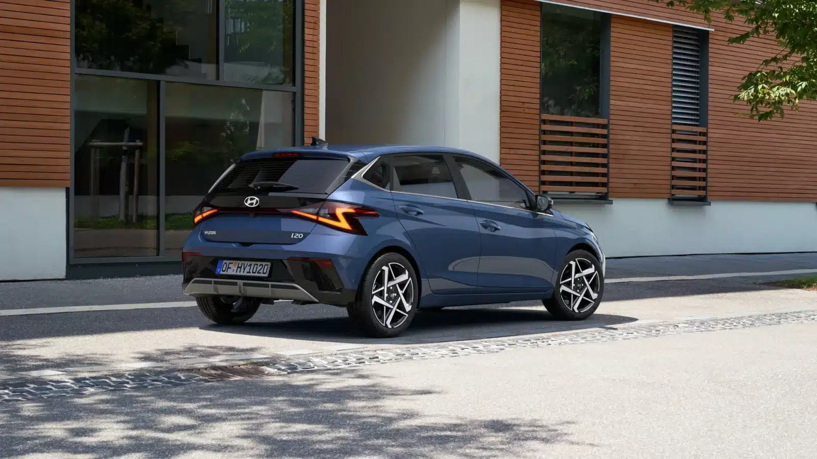 Ein blauer Hyundai i20 mit Fließheck ist am Straßenrand vor einem modernen Gebäude mit Holzverkleidung und großen Fenstern geparkt. Das Auto wird von der hinteren linken Seite aus betrachtet.