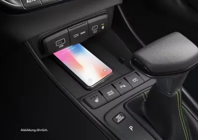 Ein Smartphone liegt auf einem kabellosen Ladepad in der Mittelkonsole eines Autos, in der Nähe eines Schalthebels und mehrerer Bedientasten. USB-, 12-V- und USB-C-Anschlüsse sind auch über der Ladefläche sichtbar.