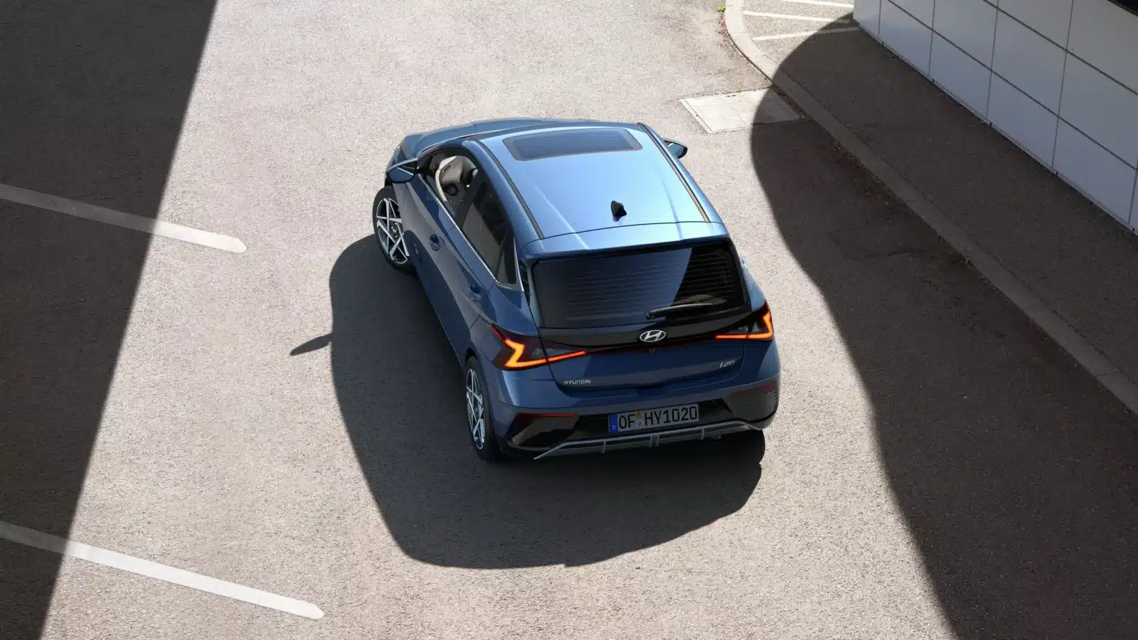 Ein blauer Hyundai i20 mit Fließheck ist auf einer gepflasterten Fläche im Halbschatten geparkt, von oben und von hinten gesehen. Das Auto hat markante Rückleuchten und ein europäisches Nummernschild.