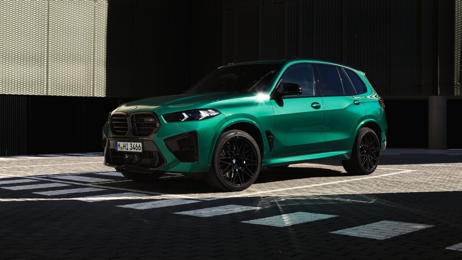 Ein metallicgrüner BMW-Geländewagen parkt auf einem gemusterten Bürgersteig vor einem modernen Gebäude, wobei geometrische Schatten und Sonnenlicht das Fahrzeug hervorheben.
