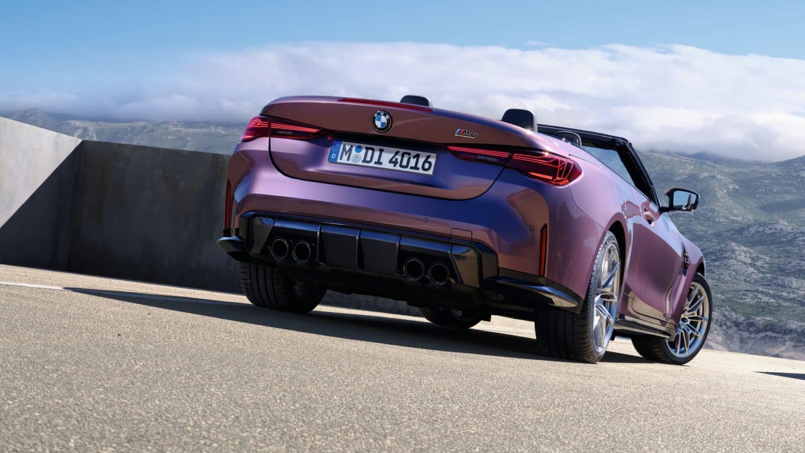 Ein metallisch lilafarbenes BMW Cabrio ist auf einer gepflasterten Fläche in der Nähe einer Betonwand geparkt, mit Bergen und einem teilweise bewölkten Himmel im Hintergrund. Das Auto wird von der hinteren Fahrerseite aus betrachtet.