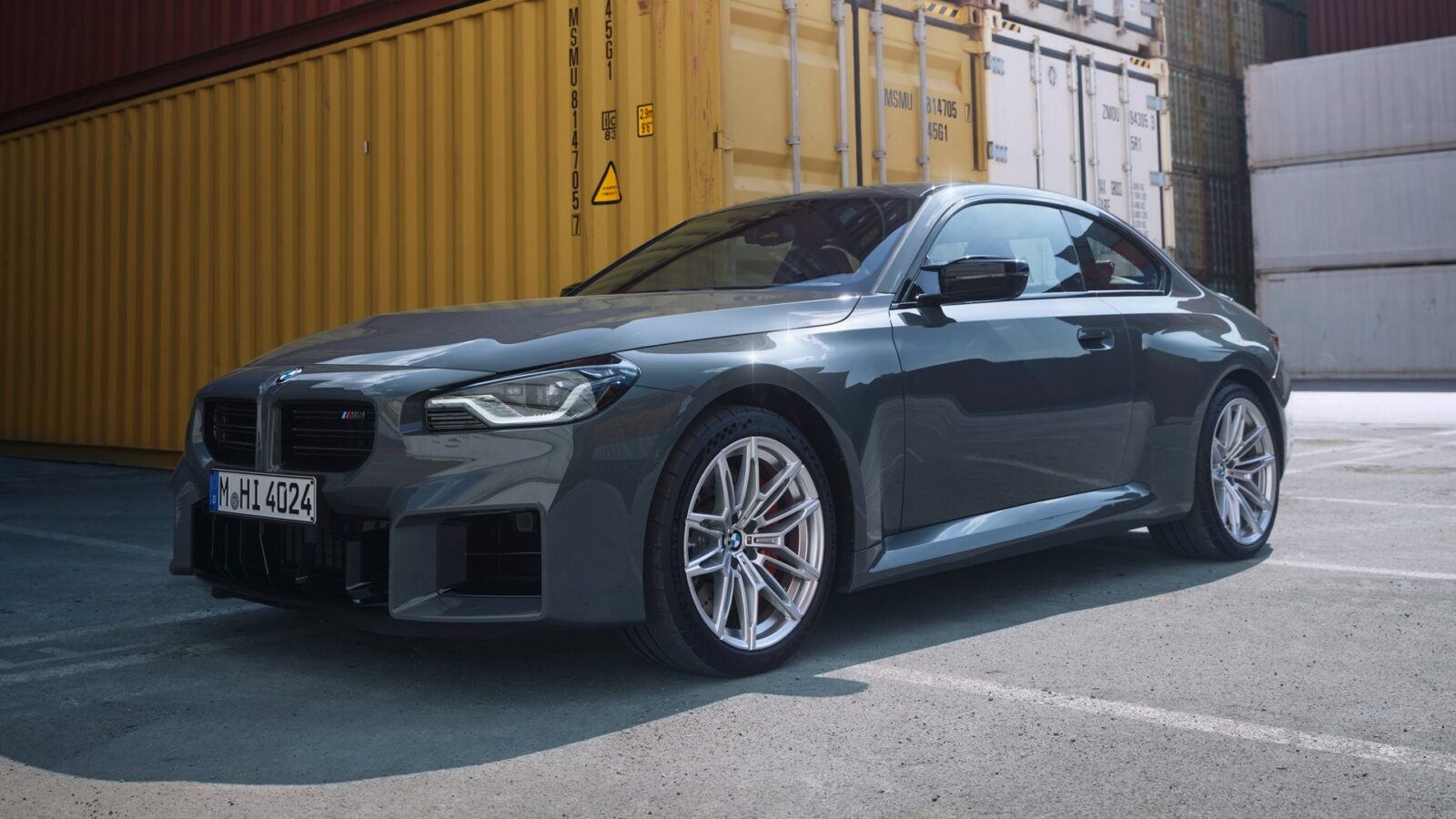 Ein graues BMW-Sportcoupé ist an einem sonnigen Tag auf einer gepflasterten Fläche vor gestapelten gelben und weißen Schiffscontainern geparkt.