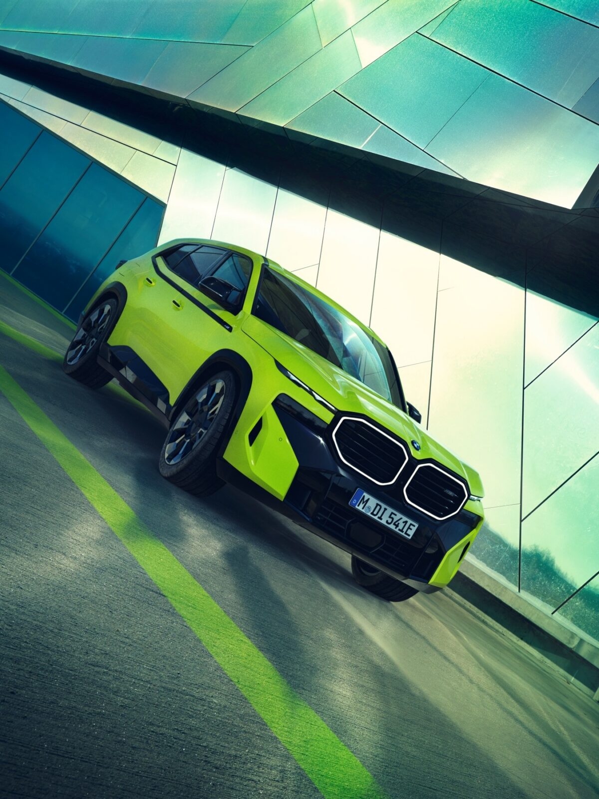 Ein leuchtend gelber BMW XM, geparkt auf einer modernen, kantigen Betonfläche mit einem Gebäude aus Glas und Metall im Hintergrund, aus einem niedrigen Frontalwinkel betrachtet.