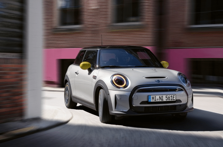 Ein silberner Mini Cooper mit gelben Seitenspiegeln fährt auf einer Stadtstraße an einem Gebäude mit rosa Backsteinwänden vorbei.